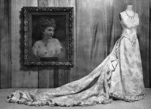 Black and white image of Elizabeth McCort Tabor's wedding dress on display at the Colorado State Historical Society. The dress is displayed on an old-fashioned manikin beside a portrait of Elizabeth McCort Tabor.
