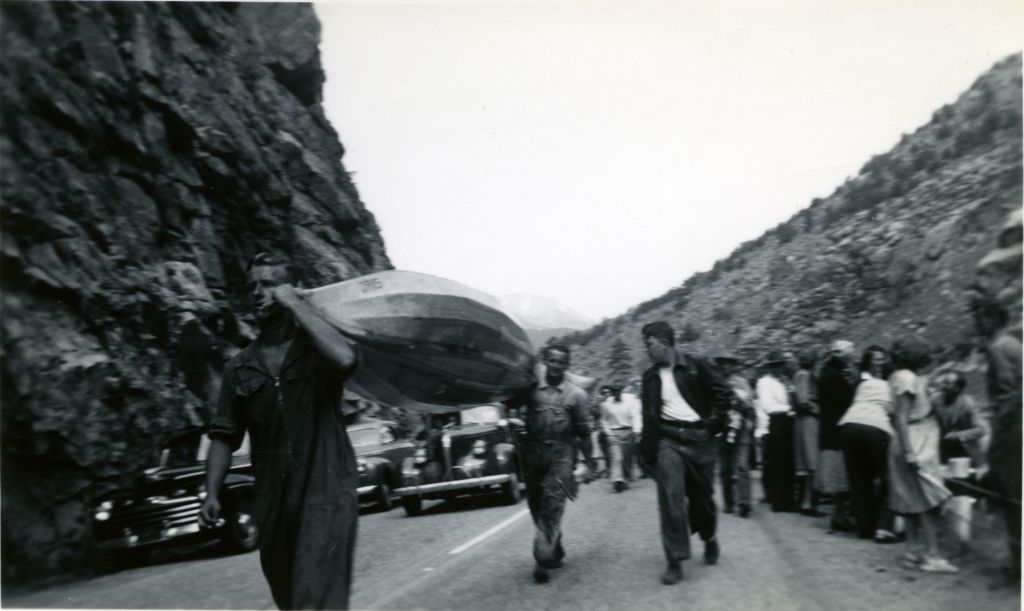 Black and white photograph of two men carrying a boat on a road beside a cliff. The men are followed by a small crowd.