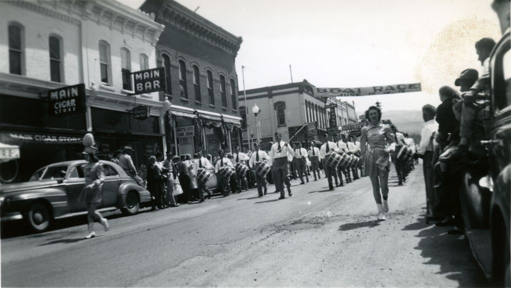 Black and white photograph of two women walking in a parade in downtown Salida, Colorado. They are followed by a group of drummers. Spectators line the street, and historic buildings for a backdrop.