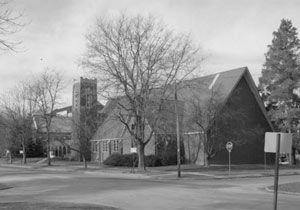 A black and white photo of the church with a large gabled roof and square steeple in the back. There are several leafless trees and an evergreen on the right.