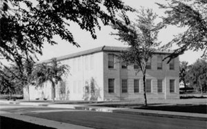 A black and white photo of the building from an angle with tall windows on the facade and trees framing the shot.