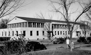 A photo of the building in black and white with light bottom half and darker top half with gable roof above. In the foreground stand some leafless trees.