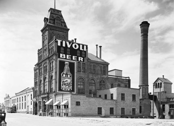 View of the Tivoli-Union Brewery and offices at 10th (Tenth) and Larimer Streets in the Auraria neighborhood of Denver, Colorado. The four-story brick building has arched windows, decorative brick work, canvas awnings and a tower with a mansard roof. A sign reads: "Tivoli Beer." Shows smokestacks, chimneys, an automobile, truck and fire hydrant.