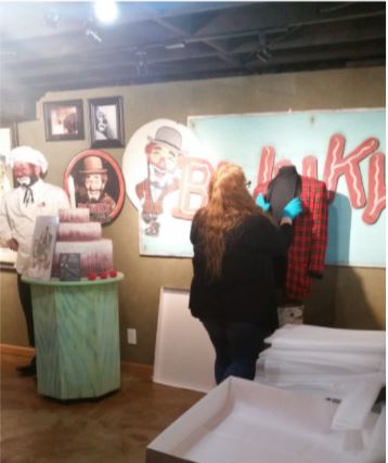 Photo of Amy Nilius, standing before a mannequin upon which Blinky the Clown's red plaid jacket hangs. She is carefully straightening it out to pack it for transportation to Denver. She is standing amidst several Blinky-related photos and memorabilia.