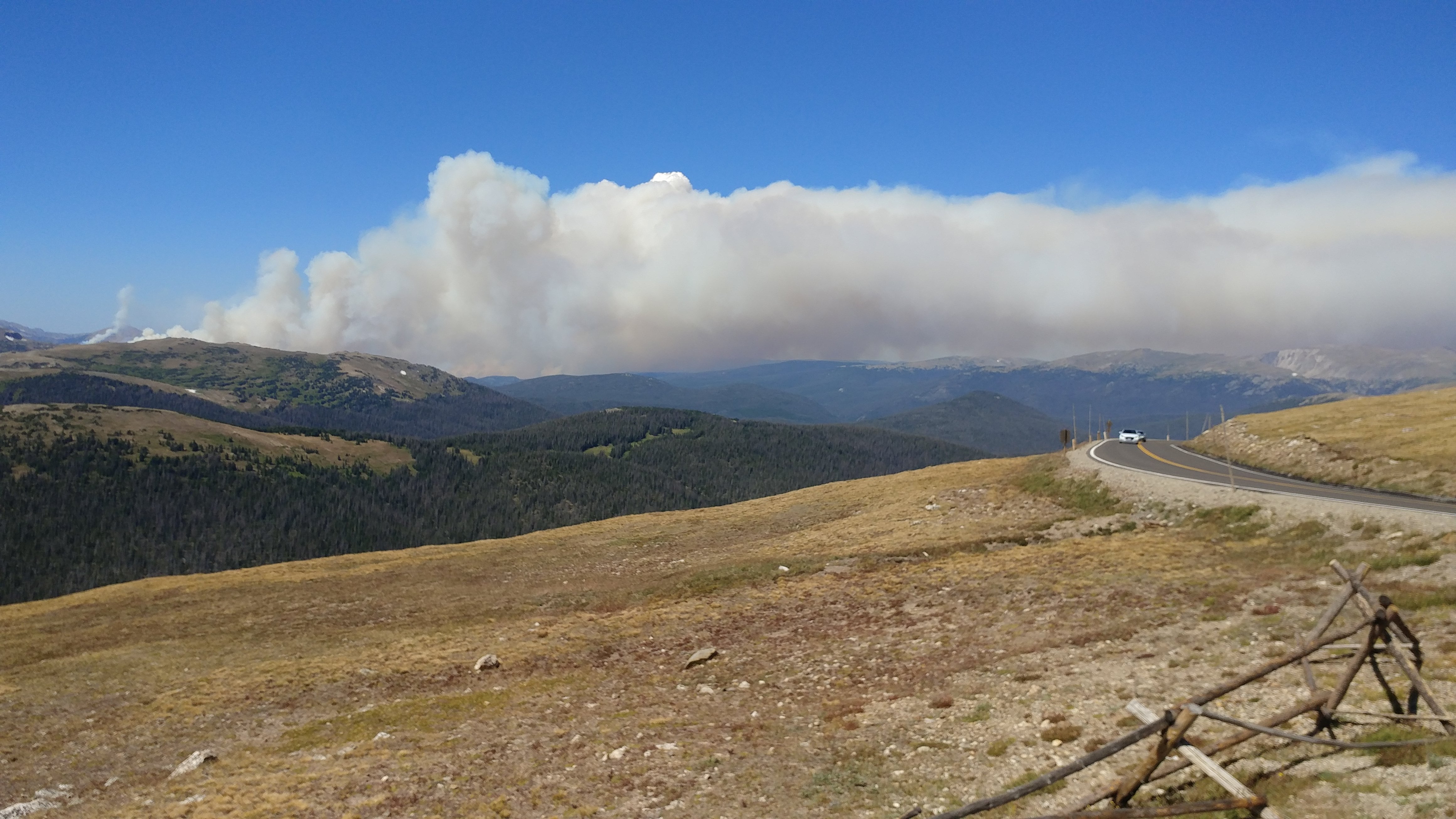 Photo of a wildfire raging in the distance along Cameron Pass. Orange flames are dancing across the top of the pass, barely visible along the horizon, while orange and gray smoke blow across the blue sky.