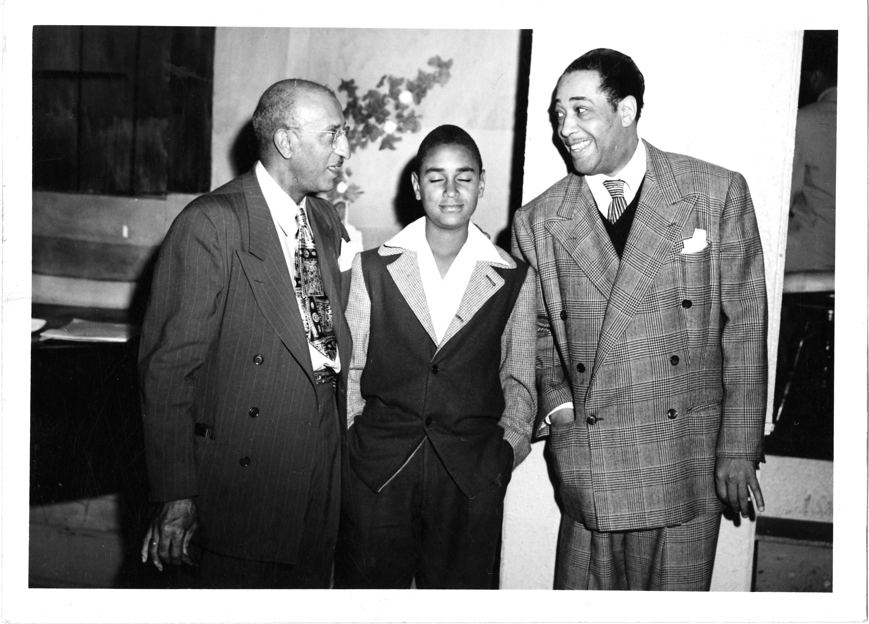 Dr. Clarence Holmes, William Holmes, and Duke Ellington stand in conversation, location is unknown. All three men are dressed in nice suits, and Duke Ellington is looking at Doctor Holmes and, by his facial expression, telling them something quite enthusiastically.