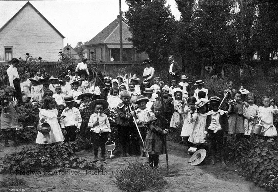 Kindergarten students at Bessemer School working in their school garden, 1902. Adults behind the class include teachers Miss Carrie Carlile, Miss Mary Williamson, and another unknown woman, most likely another teacher and Superintendent J. F. Keating.
