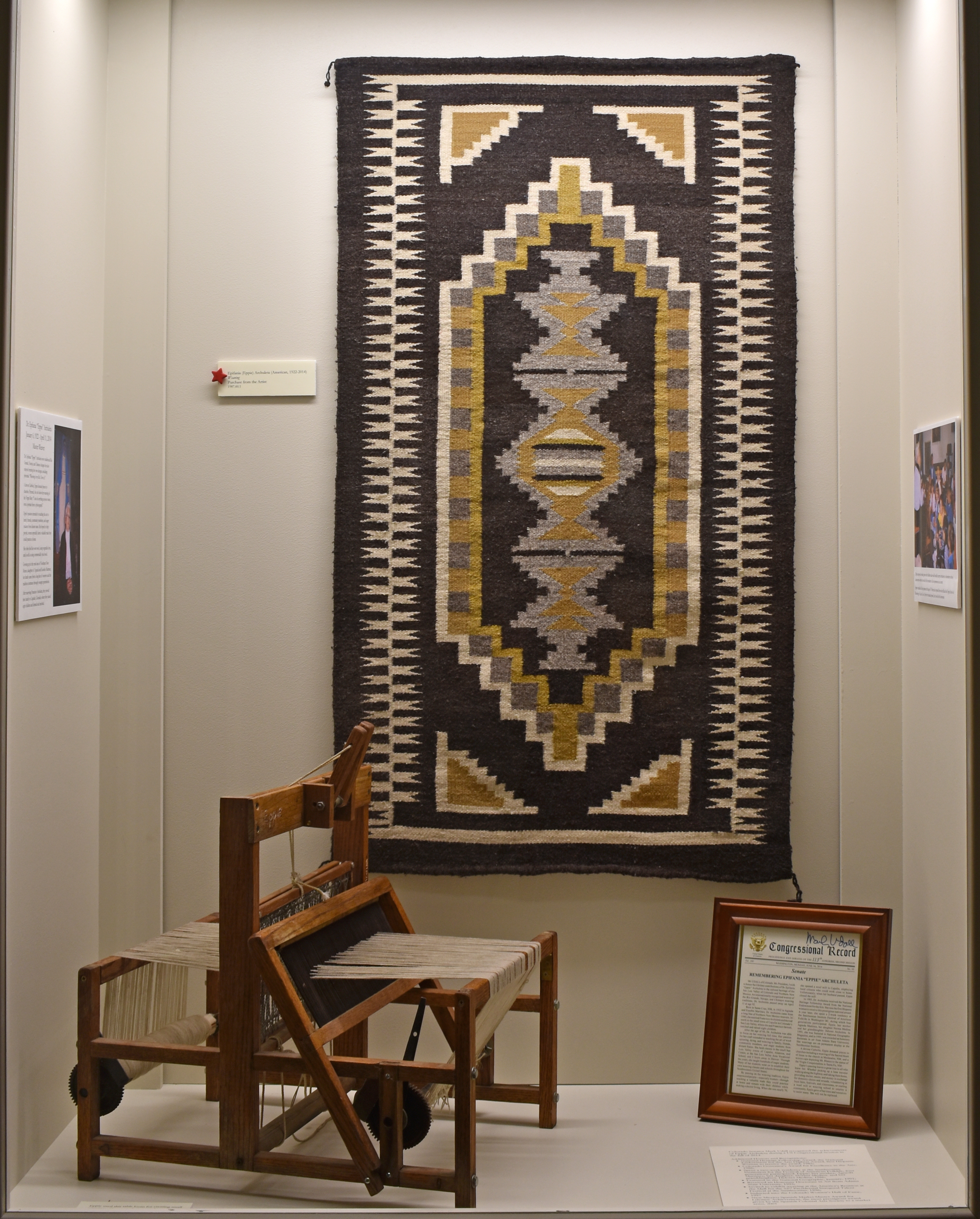 Photo of Eppie Archuleta’s weaving on display in the Adams State University Luther Bean Museum. The display includes a copy of the Congressional Record honoring Eppie for her lifetime contributions. It also displays a table loom made by Levi Medina and donated by Levi and Norma Medina.