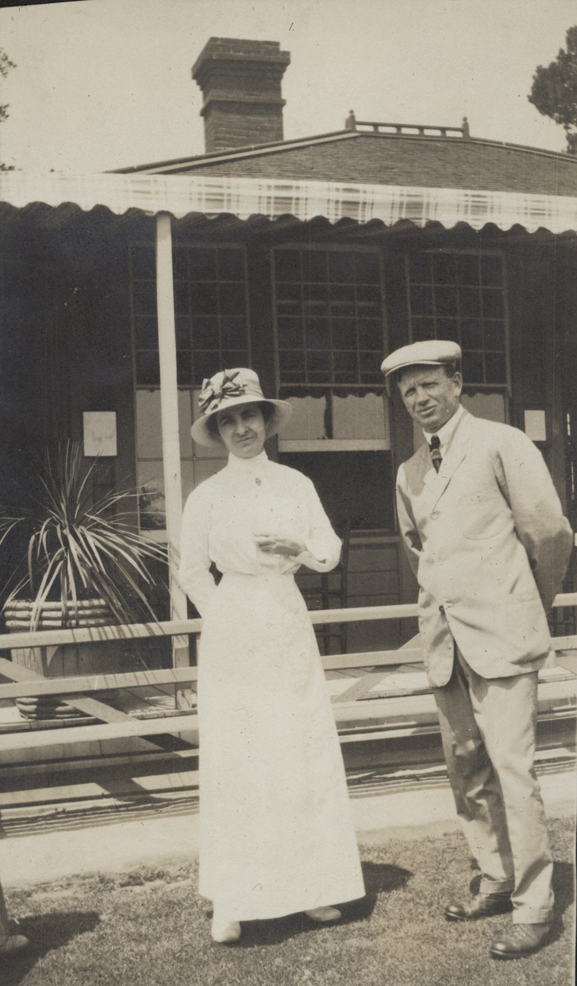 Photo of a woman and a man around the turn of the 20th century, standing in front of a two-story building. She is standing on the left, wearing a long white skirt, white shoes, a white blouse, and a wide-brimmed hat with a big bow on the front of it. He is wearing light colored trousers, leather shoes, a sport coat, and a flat cap. They are both looking at the person taking the photo. There is only a building in the background, and it is uncertain as to what the building is--there is no visible signage.