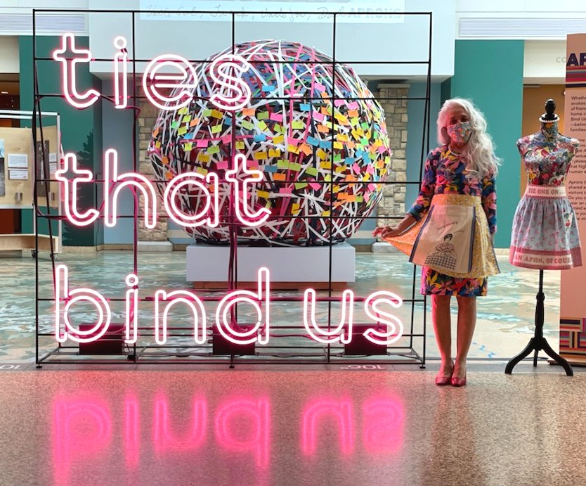 Photo of EllenAnne Geisel, standing next to a large neon sign with pink letters, that reads "ties that bind us." EllynAnne is wearing a bright floral pattern dress and a cheery yellow apron, and Covid-19 face mask. She is standing next to a mannequin that is also dressed in an apron, and all are in the atrium of the History Colorado Center in Denver.