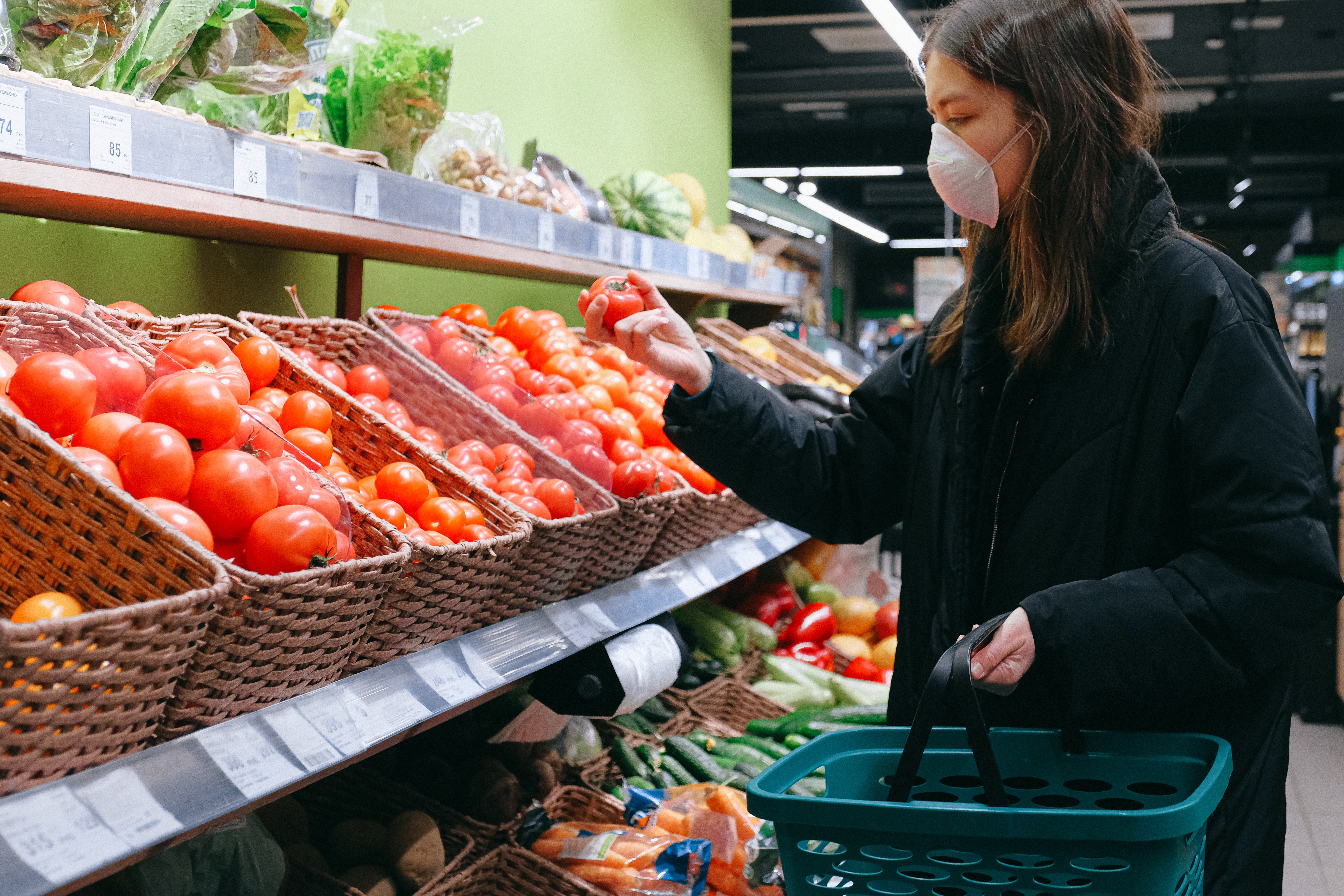 Photo of a woman grocery shopping during Covid-19 pandemic. She is choosing a tomato from the baskets of tomatoes in front of her. She is wearing a face mask and carrying a shopping basket.