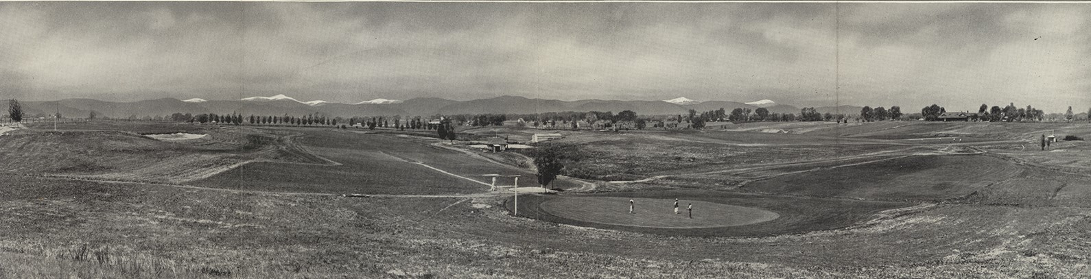 Photo of the Lakewood Country Club in 1921, taken in panoramic view. The golf course is lined with trees on the far side of the photo, and otherwise surrounded by vast open space. The snow-capped peaks of the Front Range are available on the far horizon.