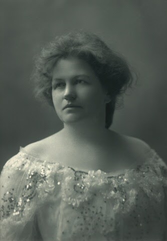 Photo of a woman posing for a portrait in a studio. The background is plain, showing off the ornate scoop neckline blouse that she is wearing. It has gathered ribbon and silver sequined details along the neckline of this fancy light-colored outfit. She doesn't wear any jewelry, and her dark hair is pulled loosely to the back of her head. She looks off to the left of the image, stoicly.
