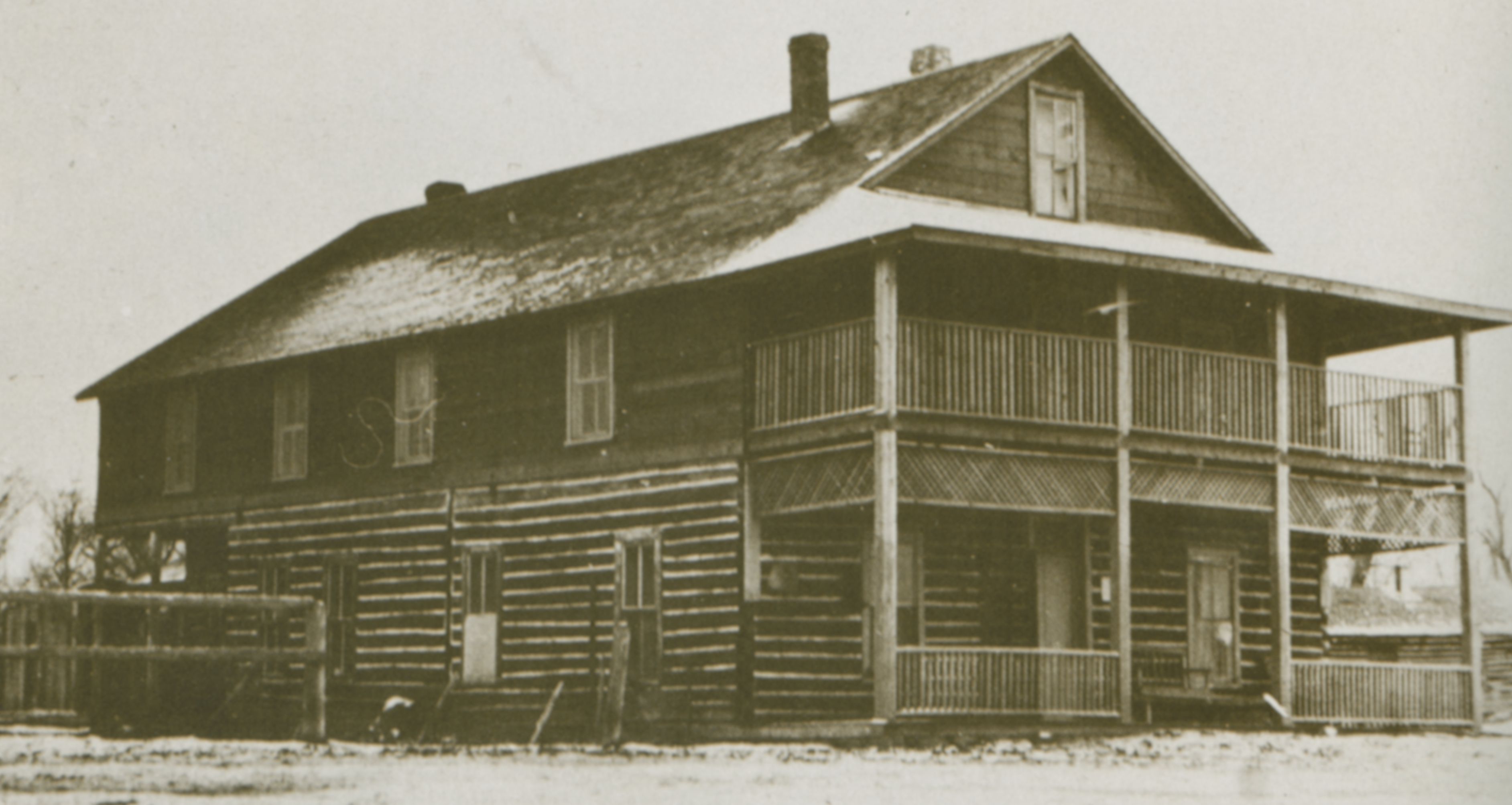 Photo of a two-story log home, with covered porches on the short end of this rectangular building. It has a simple pitched roof from which the chimney stands. The log structure has several small window on both floors and on the two sides of the home that are visible. The yard has fencing, separating land to the rear of the house from the front yard area. A dusting of snow decorates portions of the rooftop.