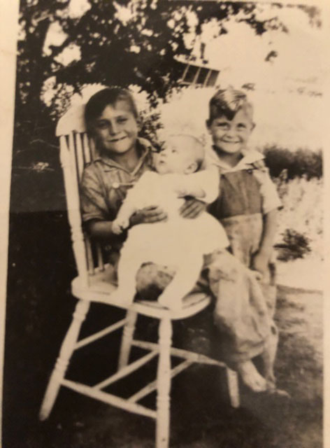 A child sits in a chair, holding a baby. Next to the chair sits another young boy.