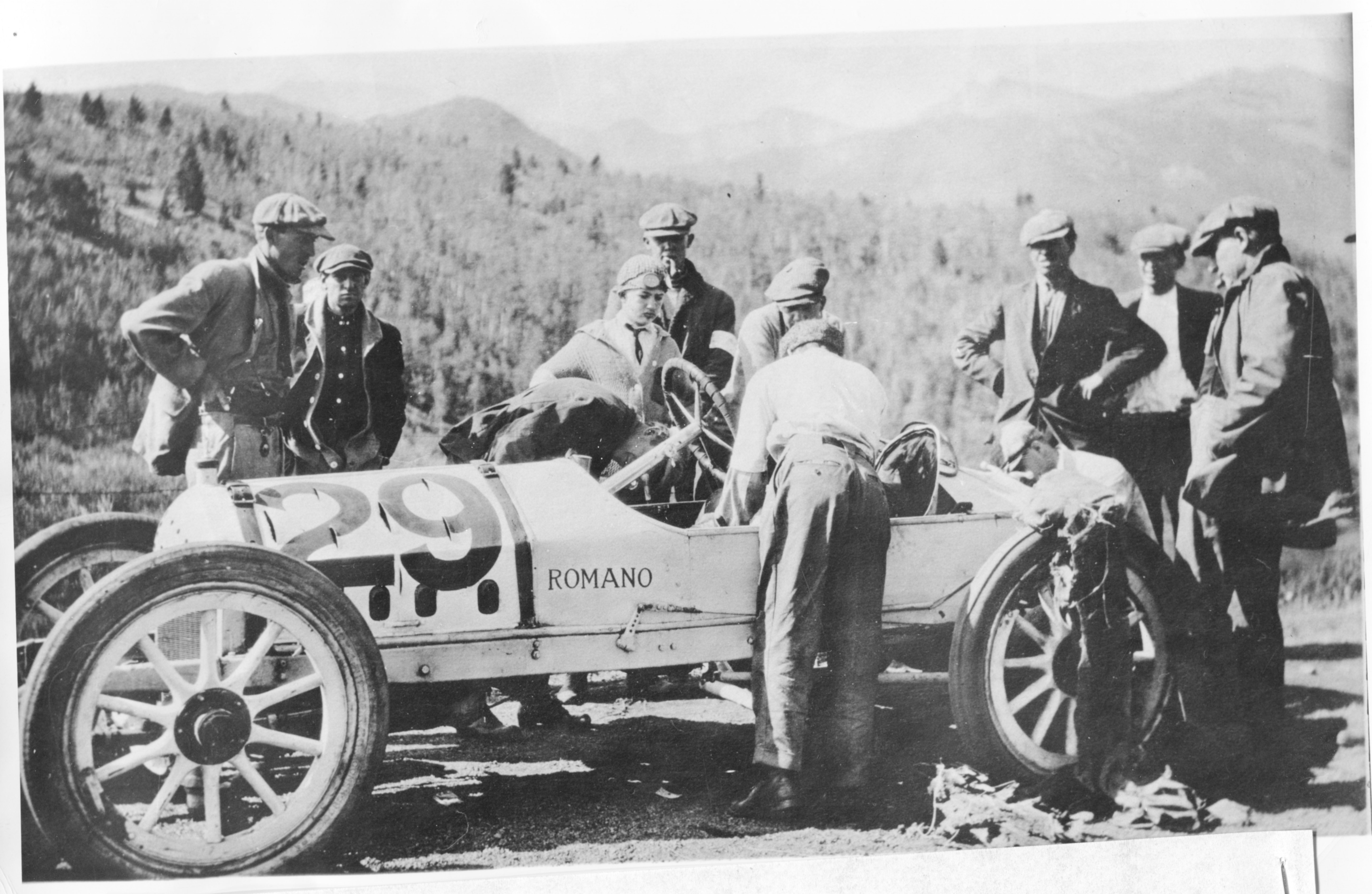 Photo of a Pikes Peak Hill Climb race car surrounded by about a dozen men in coats and flat caps, looking at the vehicle. Mountains are visible in the background. One man is leaning over the steering wheel, peering into the car. The car is open-wheeled, emblazoned with a large number 29 on the nose, and the word "Romano" in smaller letters on the driver's side.