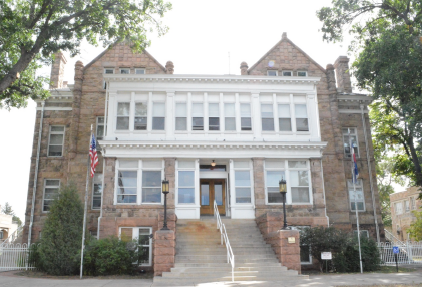 Photo of a large building that appears to be built of red sandstone bricks with white wooden window frames that surround the double front doors. A long staircase divided by a white handrail ascends to the front doors, flanked by short brick walls on each side.