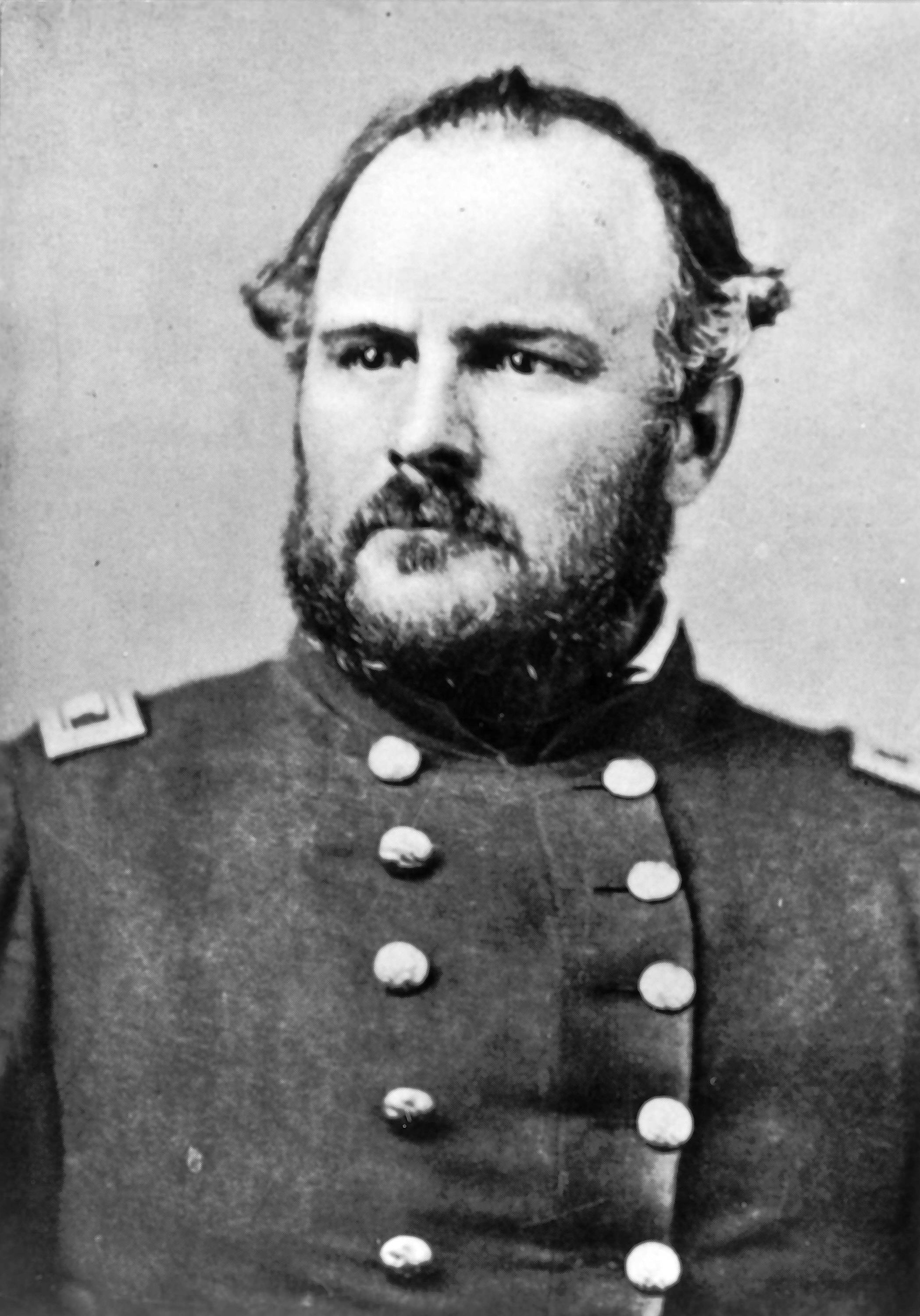 Photo fo a man in military uniform. The uniform has a double breasted placket of buttons down the center front--five large metal buttons down each side. There are patches or insignias on each shoulder of the jacket. The soldier is looking off the left as he poses for the portrait. He has a receding hairline with short curls at his temples, and a short-cropped thick beard and moustache.