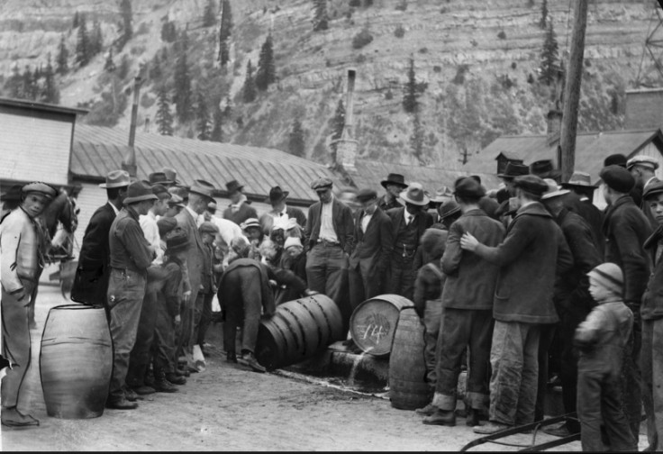A group of men in period 1920s clothing stand around a wooden barrel of alcohol while it is poured into the gutter.