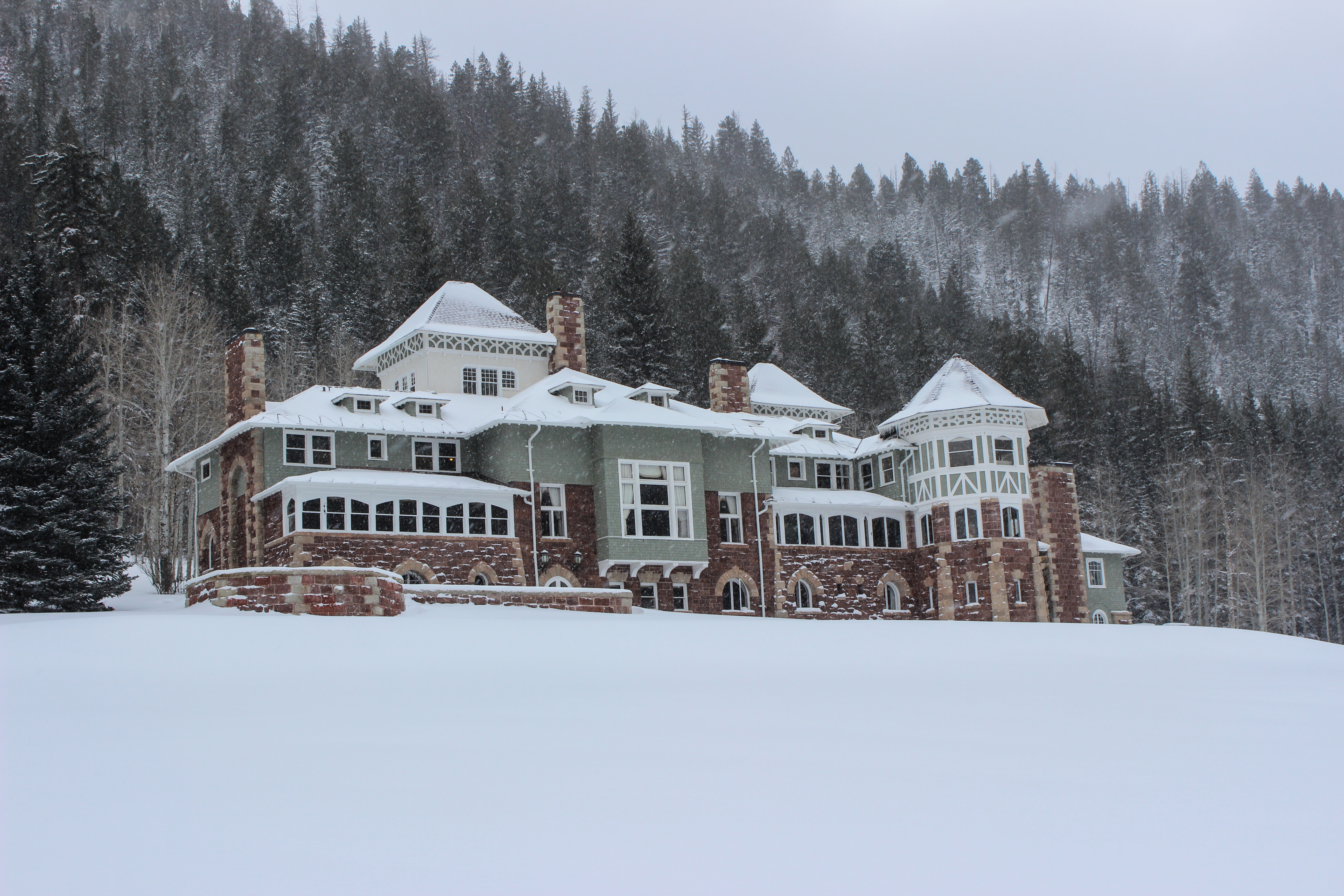 A turn of the century manor with a broad, snow-covered lawn and a dense forest behind it.