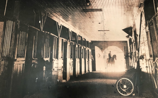 A damaged photo depicting the interior of a stable. The stable doors are open, framing the silhouette of a woman riding a horse.