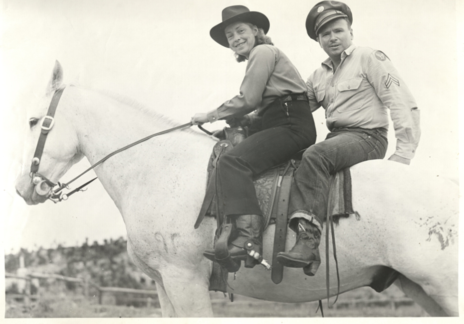 Photo of a woman and a man, riding together on a tall white horse. She wears black pants with black cowboy boots and spurs, and a long-sleeved blouse and a black cowboy hat atop her head of shoulder-length hair. He wear a military uniform shirt and hat, along with denim jeans and cowboy boots. She is holding the horse's reins as they both look at the camera and smile.