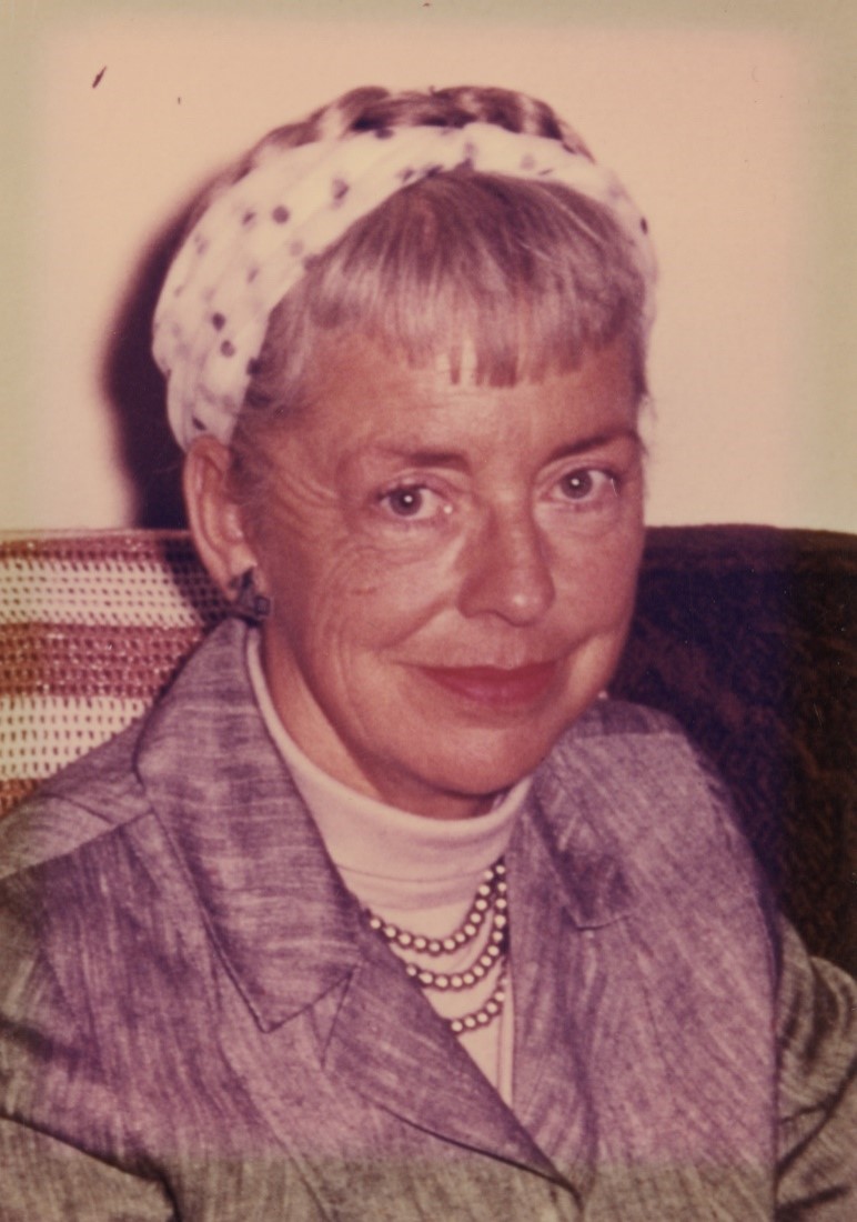 Photo of a woman, sitting for an informal portrait. She sits against a dark piece of furniture that has a gold and white striped crocheted fabric over it. She is wearing a light gray suit jacket that is buttoned up, over a white turtle neck with three strands of pearls. Her hair is braided and wrapped around her head, over which she wears a white and gray dotted head scarf. She has modest earrings and lipstick, and is smiling.