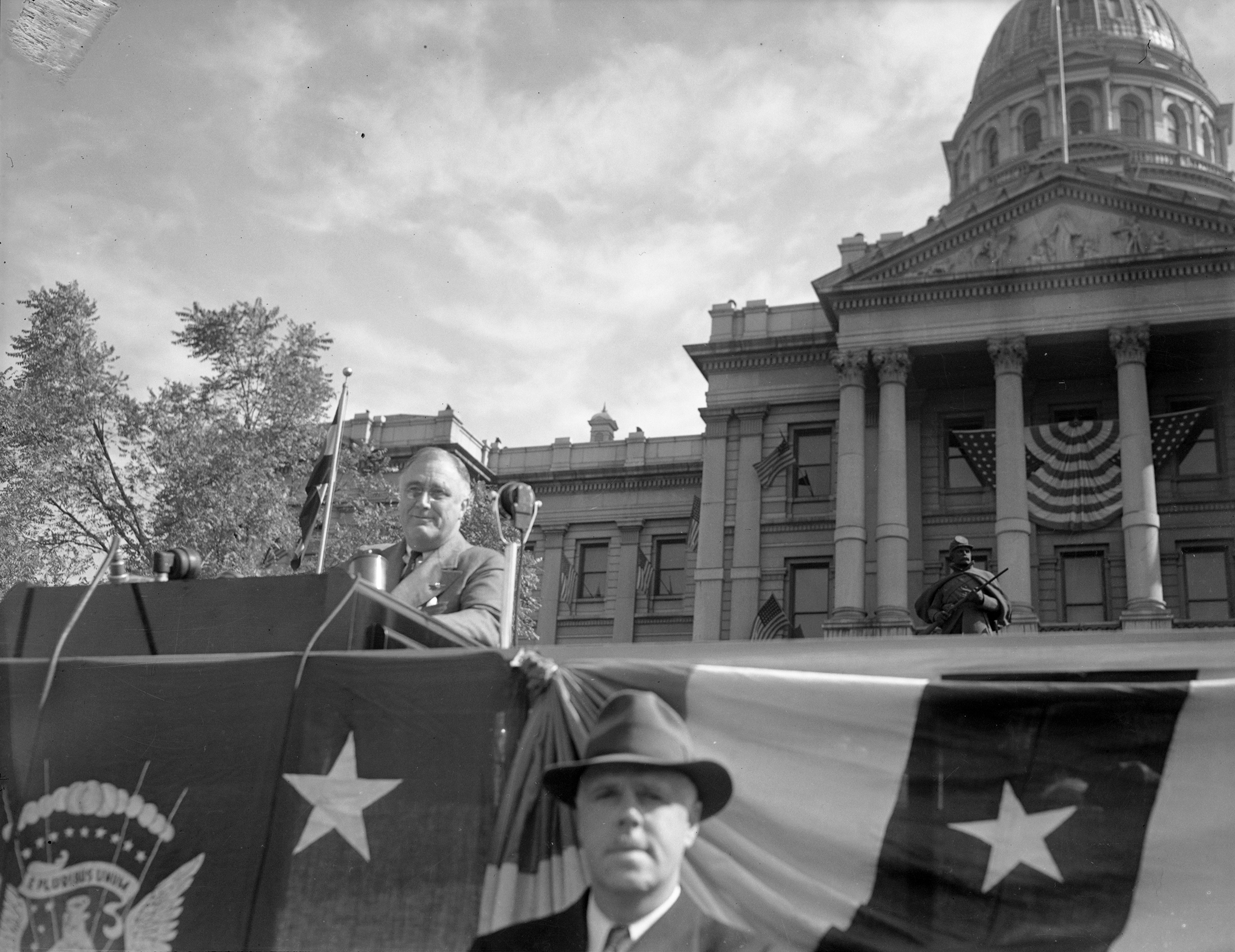 President Franklin D. Roosevelt sits behind a podium in front of the Colorado State Capitol building.