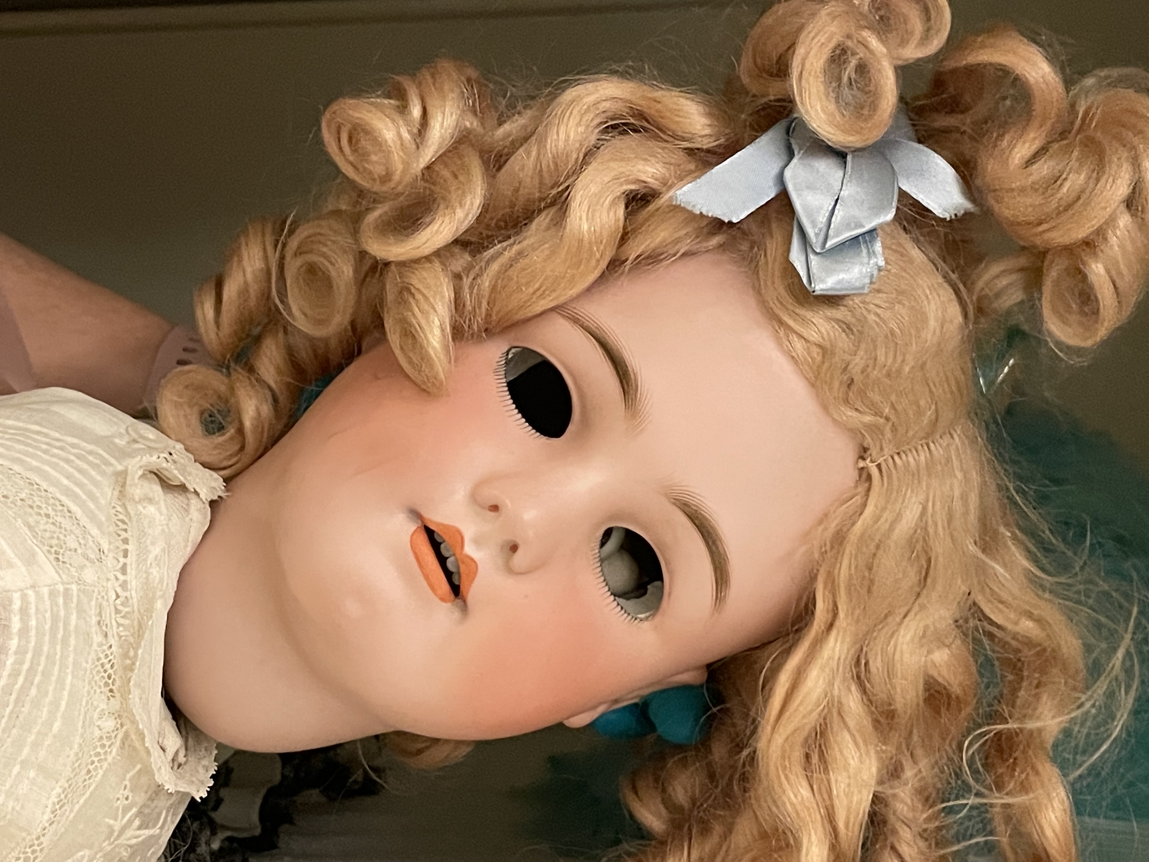 Photo of the head of a large porcelain doll, being held up by someone's left hand wearing a blue latex glove. The doll is facing toward the camera, its curly blonde hair in ringlets and some tied up with a blue ribbon on top of her head. The pink porcelain has painted-on eyebrows and orange lips, however there are no eyes or eyelids, so the viewer sees into the doll's empty head cavity.