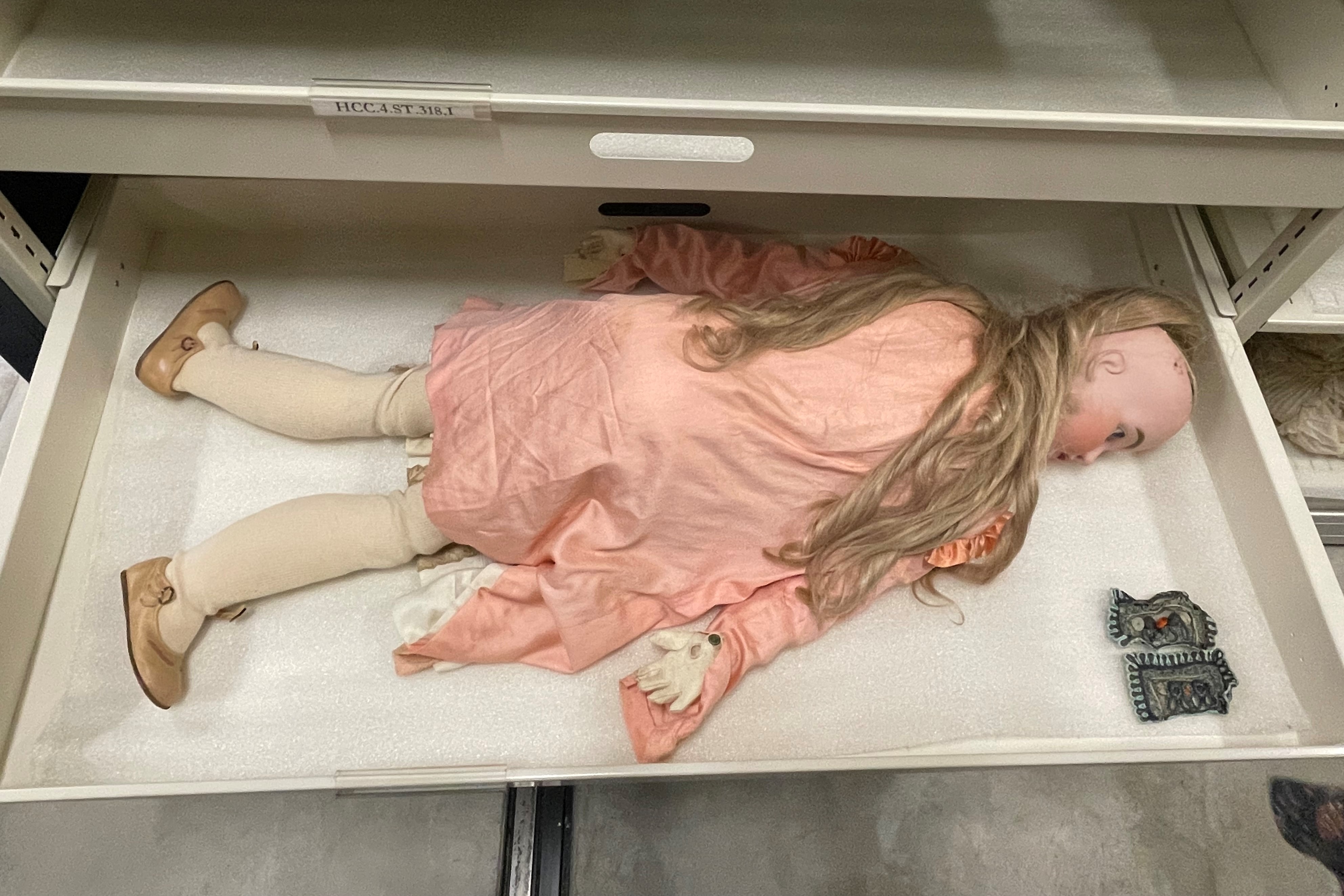 Photo of a large doll, lying facedown in a drawer in storage. The doll has long blonde hair that has slight curls at the ends, in a wig that appears to be detached from the doll head. The doll wears a pink dress with white gloves and stockings and pink buckle mary jane-style shoes.
