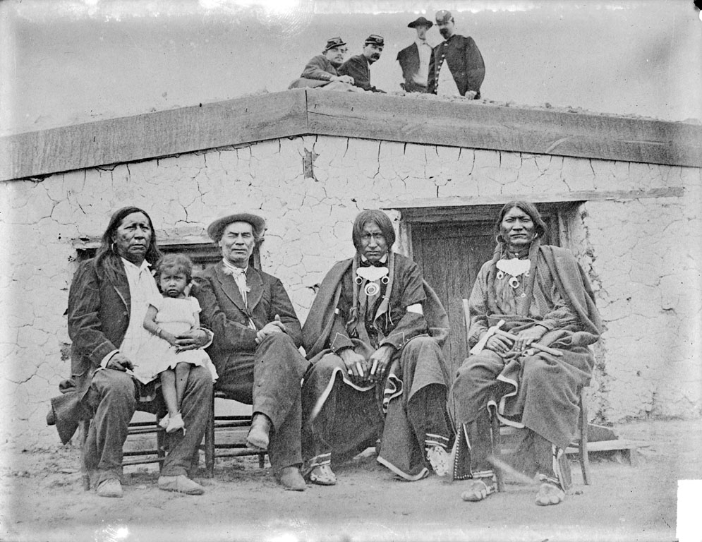 Photo of a group of people sitting in front of a one-story adobe building. A handful of people are standing on the roof, looking down toward the camera and the group seated for the portrait. Three of the four adults seated are Indigenous people, the person on the left while a toddler sits on his lap. The man to his left does not appear to be Native American, and is dressed in western-style apparel like a jacket and cap.
