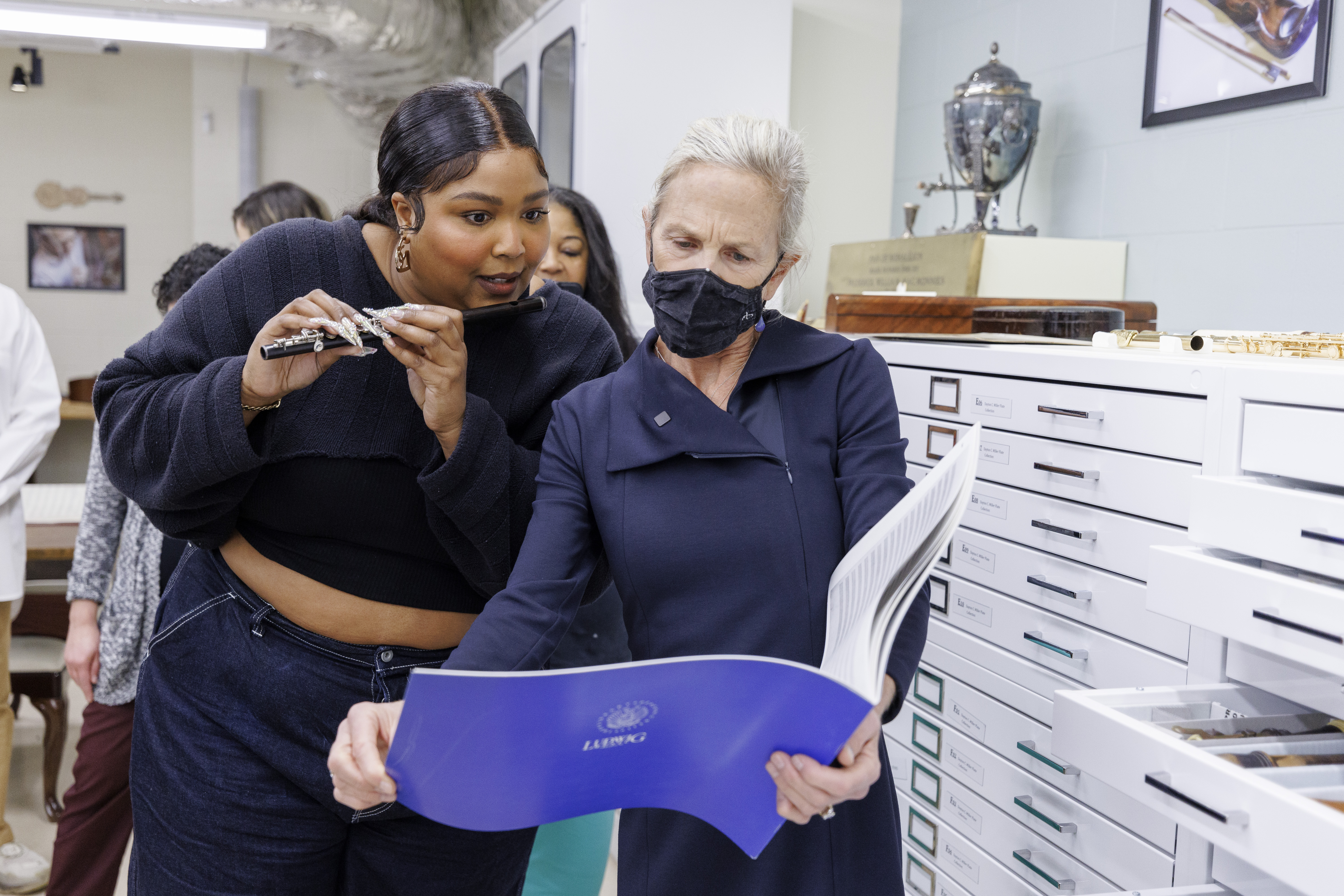 Photo of two people standing, looking at a large book with a blue cover. The Blackwoman on the left is tall and wears a navy blue sweater and jeans. She is holding a flute in both hands as if preparing to play it, looking with enthusiasm at the pages of the book. The book itself is held by the white person on the right, who is wearing a navy blue jacket and a black face mask. She has silver hair that is pulled back away from her face. They are standing in a white room, in front of a cabinet