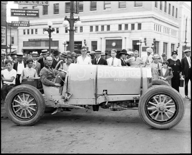 Photo of a Pierce-Arrow car which has been turned into a race car to compete in the Pikes Peak Hill Climb. The open-wheeled car is painted yellow, although that is not obvious in this historic black and white photo. An unknown man sits behind the steering wheel, as crowds of people watch him or look at the camera. The car in on display on a town street, as buildings, lamp posts, and signs are visible in the background. One sign says "Pikes Peak Cog Road" and another says "Broadmoor-Cheyenne Mountain Highway