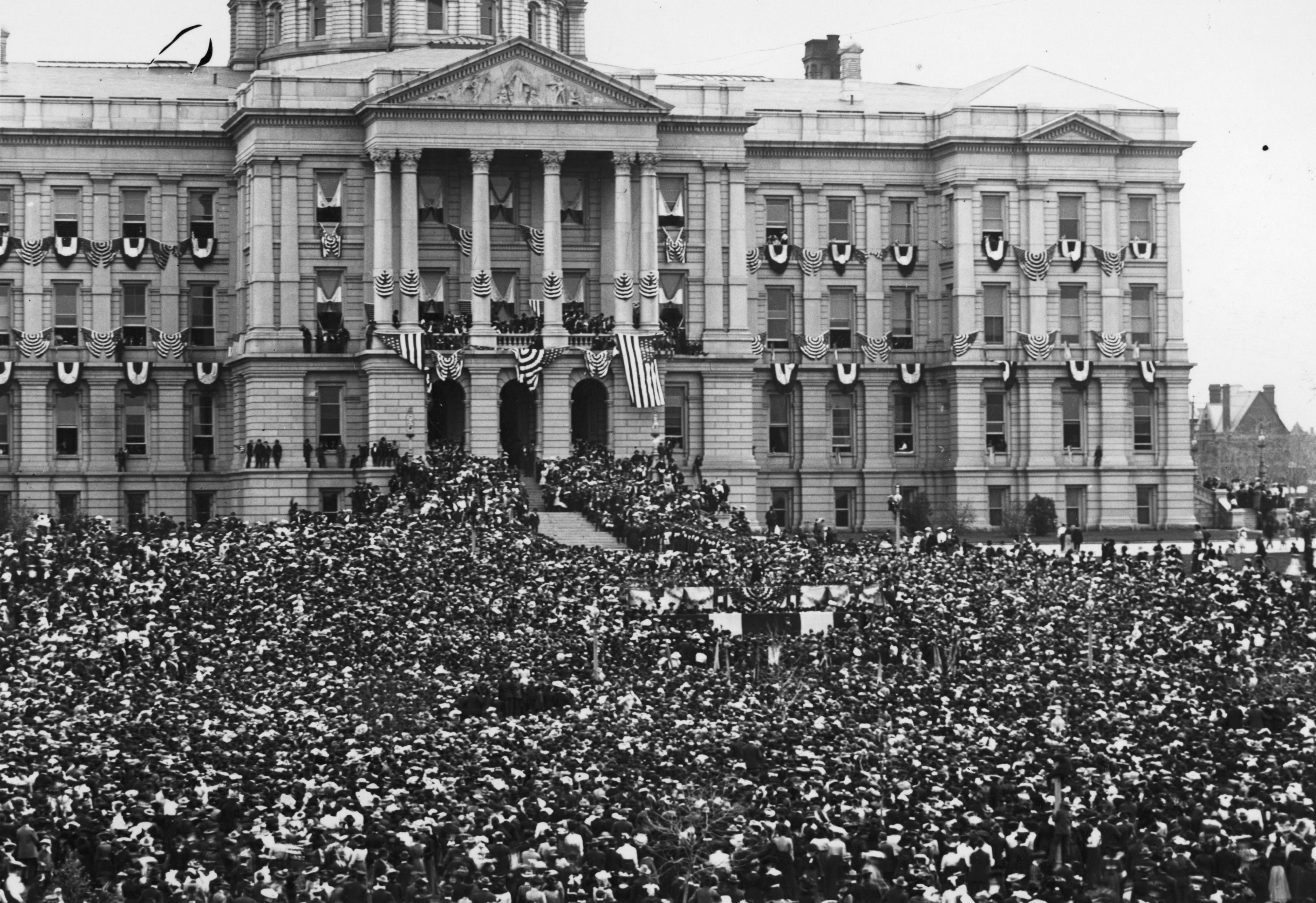 A crowd of people are gathered outside the front steps of the Colorado State Capitol building.