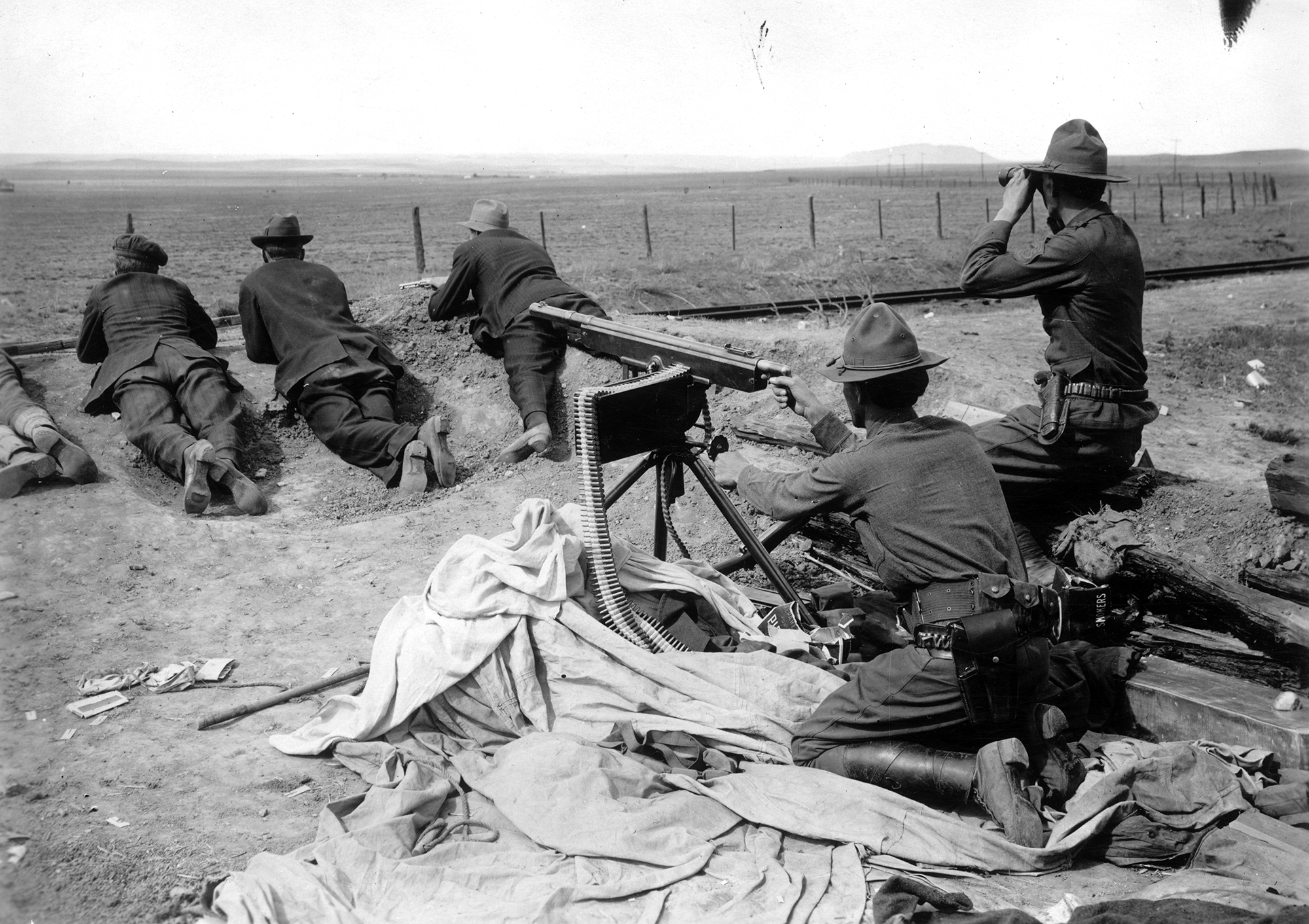 A man in Colorado National Guard uniform a machine gun nest. He is pointing it away from the camera. Another National Guardsman kneels next to him with binoculars. Three more are laying prone in front of the machine gun.