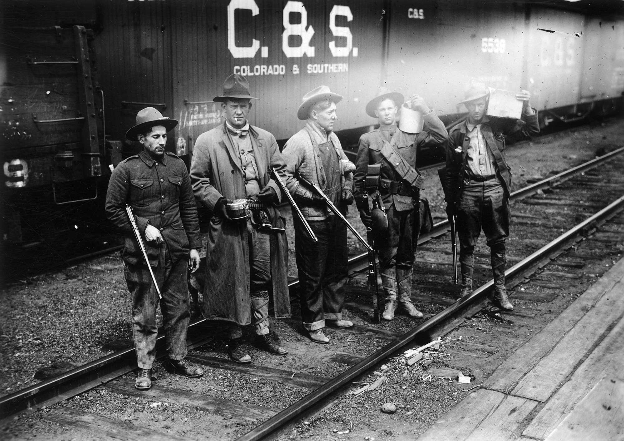 Four men standing on rail road tracks in front of a box car labelled C&S. The men are wearing hats and holding rifles.