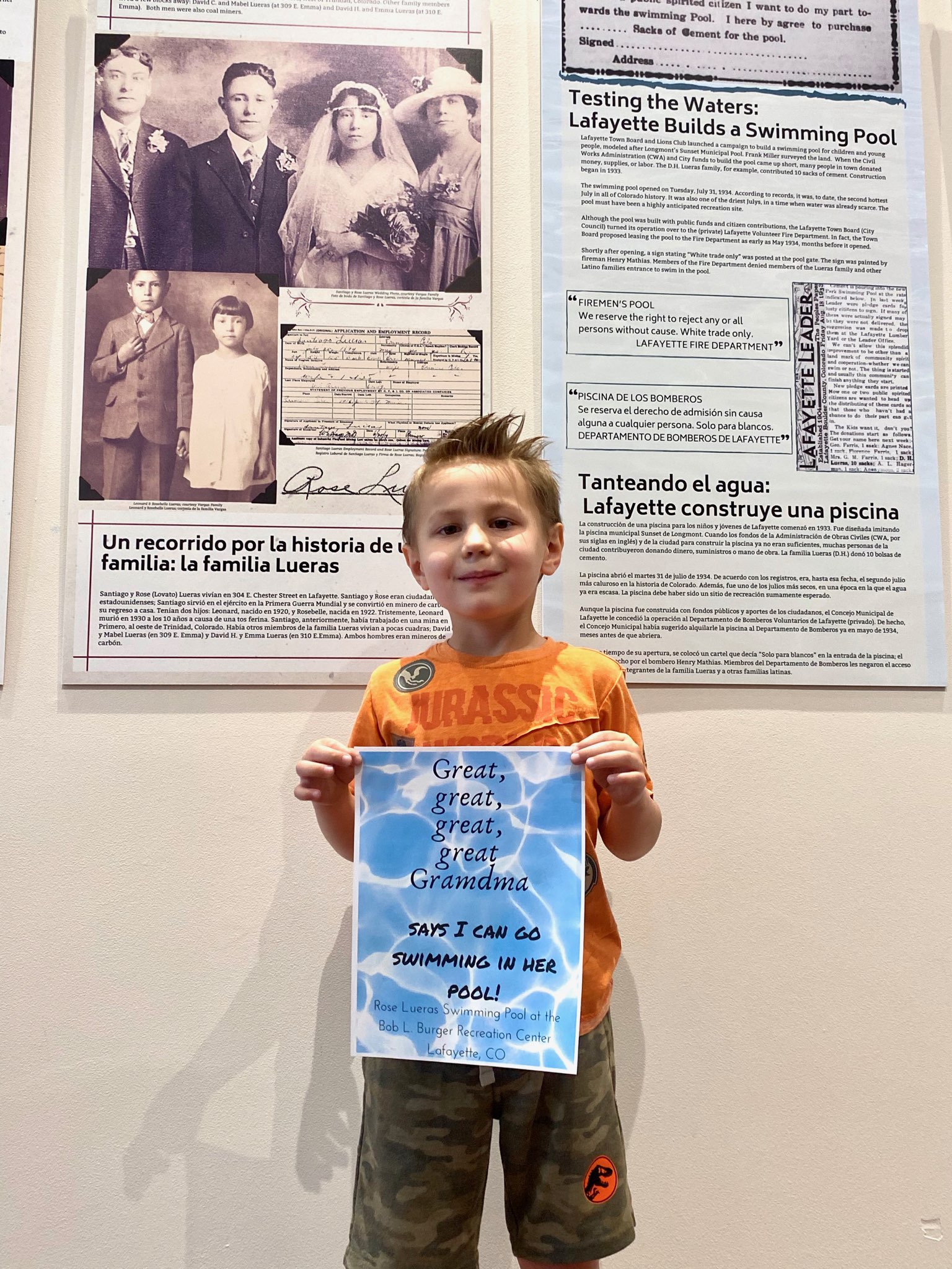 A small boy stands in front of bilingual text panels, holding up a sign which reads: Great-great-great-great-Grandma Says I Can go Swimming in Her Pool.