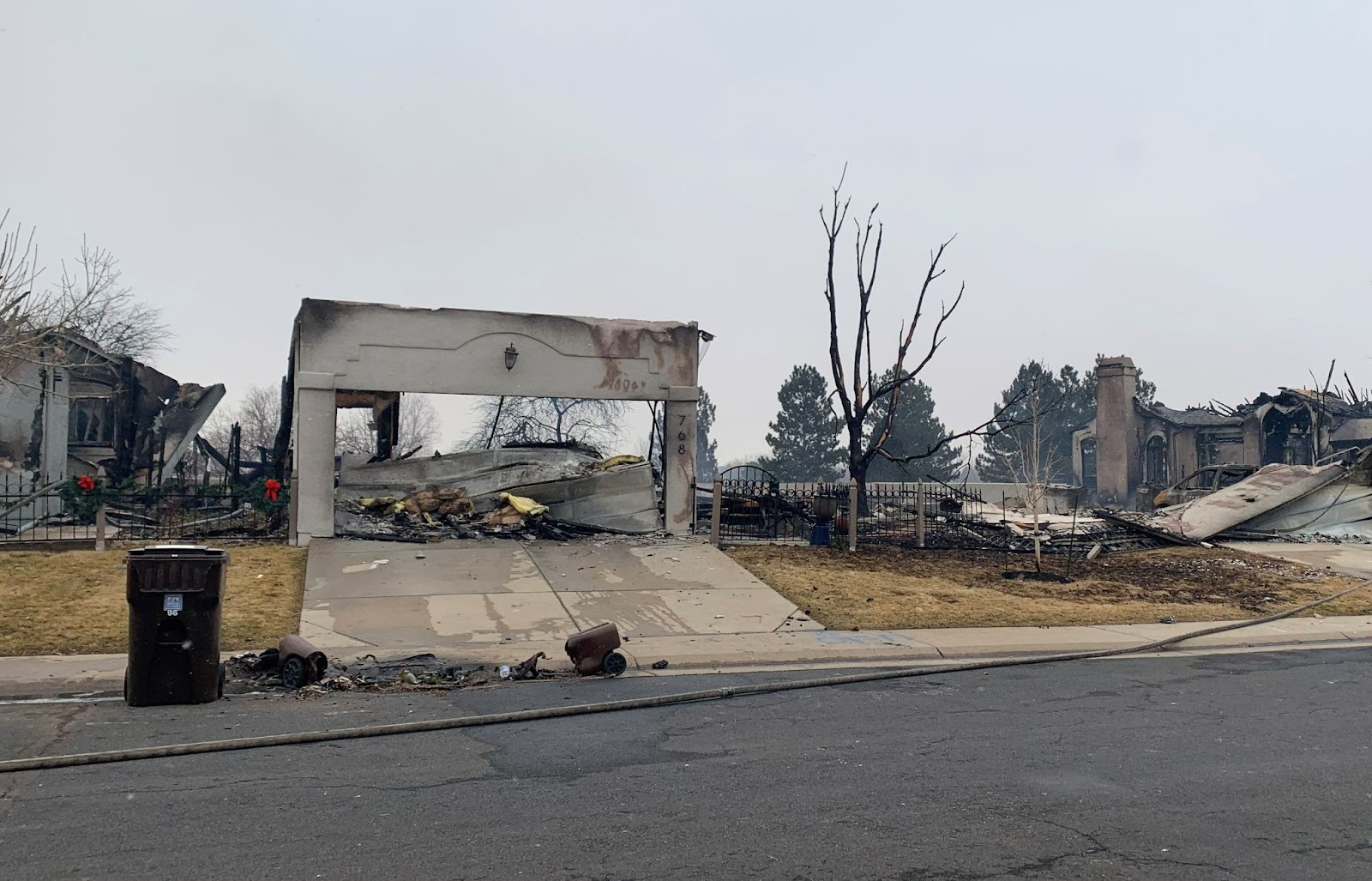 A burned house sits on a street. Only one front wall is left partially standing, and through the gaping garage door a pile of rubble is visible.