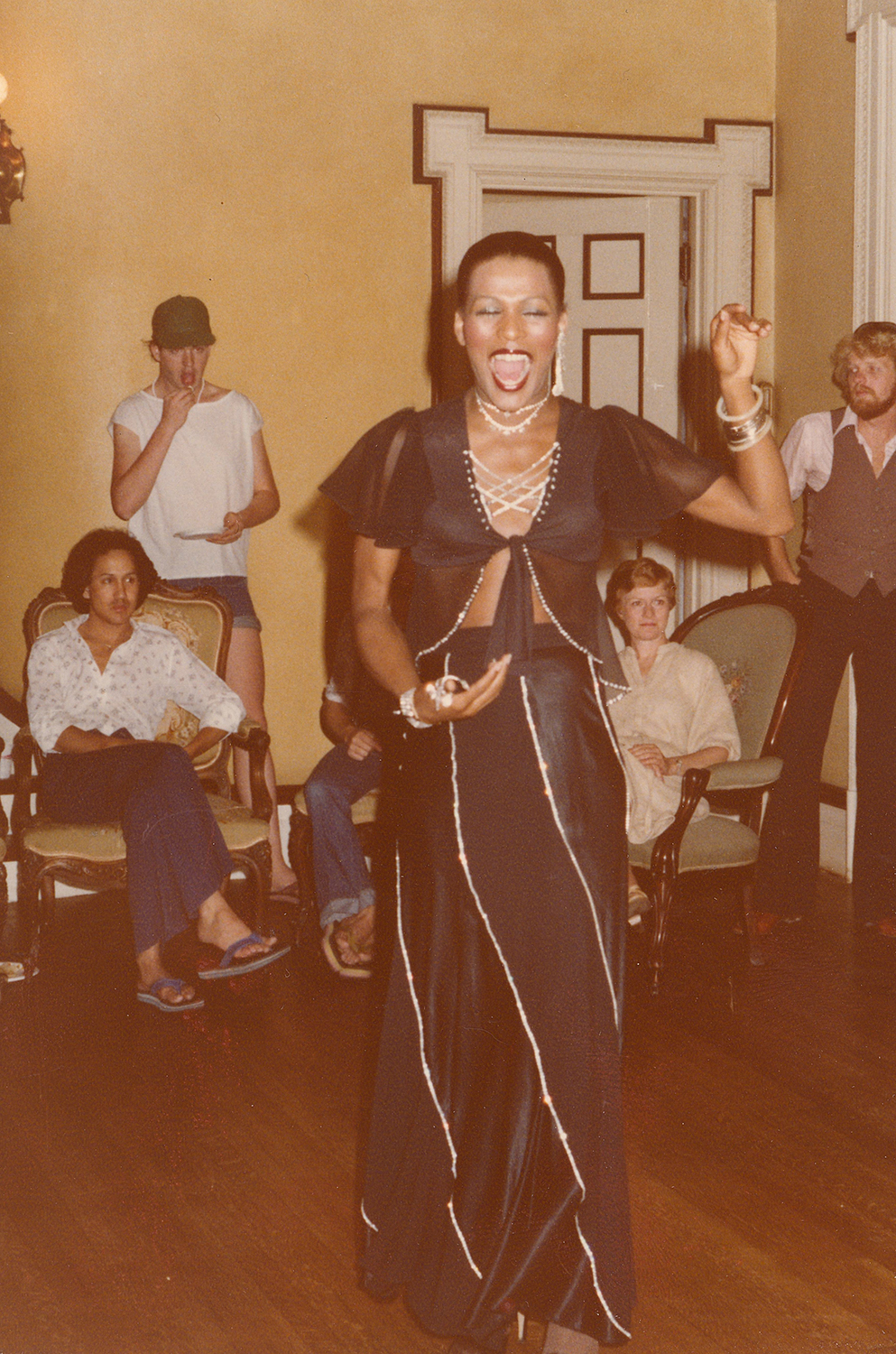 Photo of a person dancing in front of a room full of people. There are a handful of people visible behind the dancer, standing to watch and sitting in armchairs. The singer is standing on the hardwood floor, holding their right hand in front of their waist and their left hand is raised above their shoulder, eyes closed and mouth open in song. The performer wears a floor-length black striped skirt, with a black blouse that ties in the front and is embellished with sparkling crystal trim.