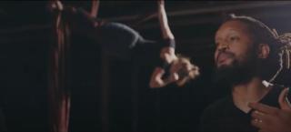 Photo of the author in the foreground of a dark and dimly-lit warehouse setting. This image captures the author reading his poem while an aerial performance artist is contorting while suspended from the ceiling with red fabric for ropes.