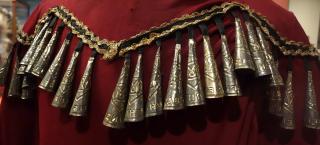 Photo of the back yoke detail of a red jingle dress. Black contrasting trim and gold rick rack in a zigzag pattern across the back shoulders of the dress, and the individual jingle pieces are attached with black fabric. The jingles themselves are each made of the lid of a metal tobacco can, and curled into a conical bell shape. The small end is affixed to the black fabric, and as the dancer moves, the jingle pieces touch to create sound.