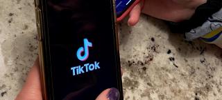 Photo of two people holding cell phones that are open to the TikTok app. One phone has the TikTok logo, and the other phone shows a photo of a man and his cat--the cat is wearing a hat and meowing.