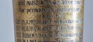 Photo closeup of the inscription on a silver-plated vase that shows some tarnish. The trophy pictured is the original presented to the winner of the Pebble Beach Golf Tournament, which began in 1920. The inscription shows that M.A.McLaughlin won the trophy in 1920, 1921, and 1923. A Robert Hunter won it in 1922.