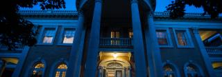 A photograph of the front facade of the Grant-Humphreys Mansion at night. The house is mostly shrouded in blue near-darkness, but lights are on inside.