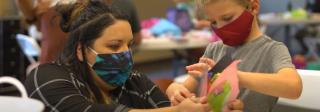 A museum staff member helps a Hands-On History student with a craft at El Pueblo History Museum. Both are wearing facemasks.
