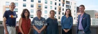 A group of six individuals stand on a balcony in Denver, with the city skyline and cranes behind them.