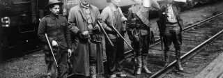 Four men standing on rail road tracks in front of a box car labelled C&S. The men are wearing hats and holding rifles.