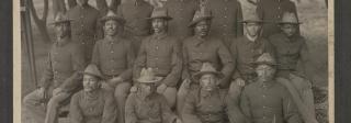 Black and white photo of soldiers in uniform standing or sitting in rows in front of trees.