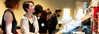 A group of people standing in front of display cases at our exhibition, Zoom In. 2 women with short hair in black dresses speak.