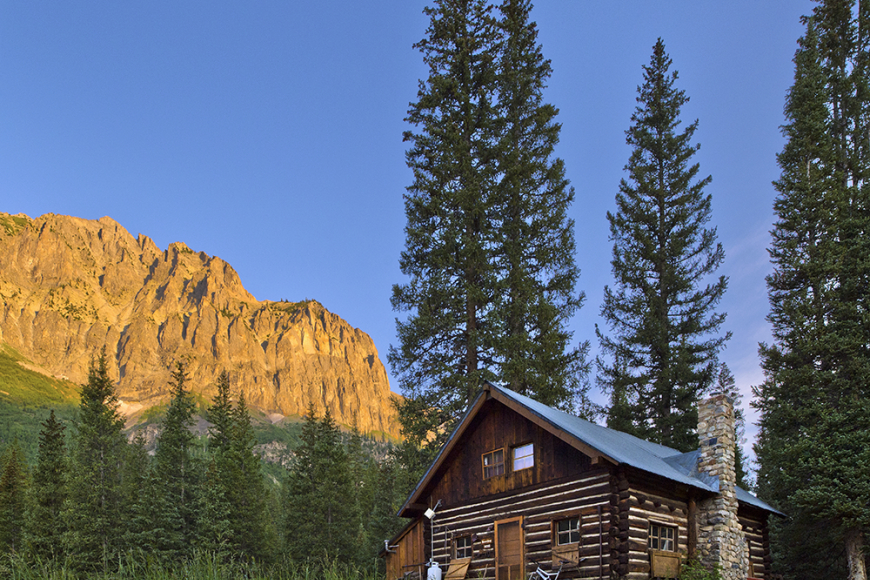 Barclay Cabin sits nestled among tall pine trees. The building is a small log structure in good condition, with a steepled tile roof. Behind it a large geological formation is visible, illuminated by sunlight.